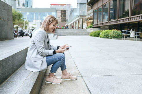 Lächelnde Frau sitzt auf einer Treppe in der Stadt und benutzt ein Mobiltelefon - KIJF02823