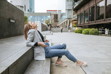 Glückliche Frau sitzt auf einer Treppe in der Stadt und lehnt sich mit ihrem Handy zurück - KIJF02824