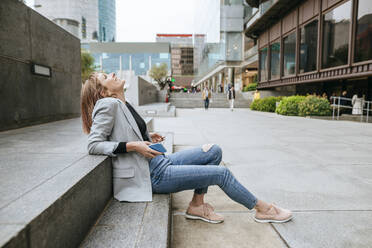 Happy woman sitting on stairs in the city with cell phone leaning back - KIJF02824