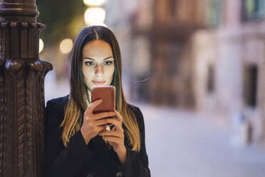 Porträt einer jungen Frau, die sich abends an einen Laternenmast lehnt und auf ihr Handy schaut - DLTSF00284
