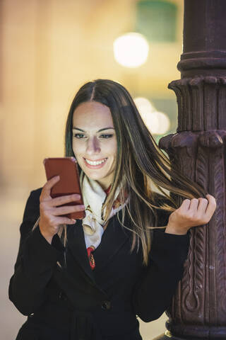 Porträt einer lächelnden jungen Frau, die sich abends an einen Laternenmast lehnt und auf ihr Handy schaut, lizenzfreies Stockfoto