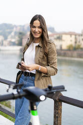 Junge Frau mietet einen E-Scooter und hält ihr Smartphone in Verona, Italien - GIOF07724