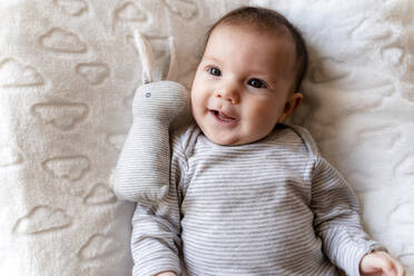 Baby girl lying on a white blanket with clouds and a rabbit toy - GEMF03301