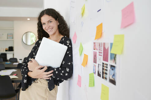 Portait of smiling young businesswoman leaning against a wall full of sticky notes in an office - IGGF01466