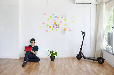 Junger Geschäftsmann, der Kopfhörer trägt und ein digitales Tablet benutzt, während er in einem modernen Büro auf dem Boden sitzt - IGGF01469