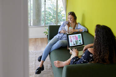 Zwei junge Geschäftsfrauen sitzen auf einer Couch in einem modernen Büro - IGGF01476