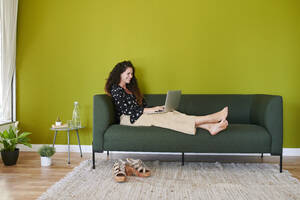 Lächelnde junge Geschäftsfrau mit Laptop auf der Couch in einem modernen Büro - IGGF01477