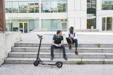 Two casual businessmen talking on stairs in the city next to e-scooter - IGGF01493