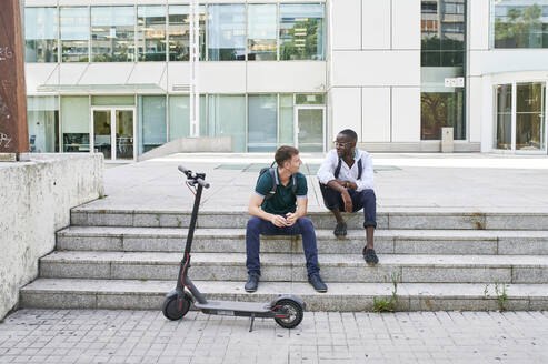 Zwei lässige Geschäftsleute unterhalten sich auf einer Treppe in der Stadt neben einem E-Scooter - IGGF01493