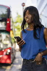 Afroamerikanerin mit Smartphone an einer Bushaltestelle in London, Vereinigtes Königreich - MAUF03067