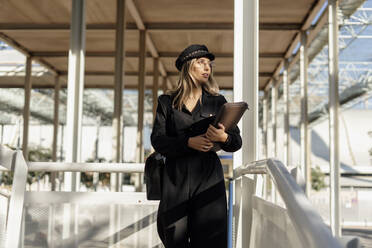 Junge blonde Geschäftsfrau mit schwarzer Seemannsmütze und Laptoptasche in der Hand - ERRF02072