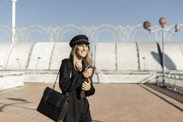 Junge blonde Geschäftsfrau mit schwarzer Seemannsmütze und Laptoptasche in der Hand - ERRF02073