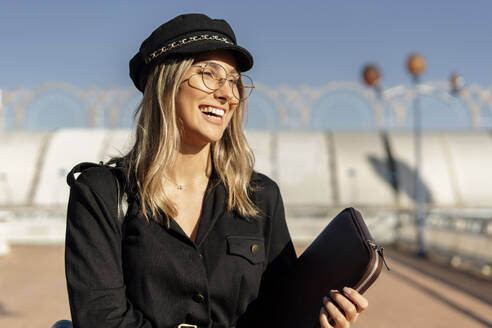 Junge blonde lachende Geschäftsfrau mit schwarzer Matrosenmütze und Laptoptasche in der Hand - ERRF02074