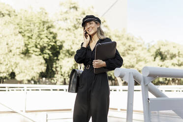 Junge blonde Geschäftsfrau mit schwarzem Hut, Laptoptasche und Smartphone in der Hand - ERRF02076