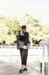 Young blond businesswoman wearing black hat and holding laptop bag and using smartphone - ERRF02077