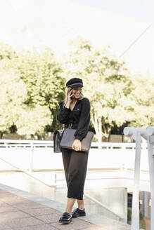 Junge blonde Geschäftsfrau mit schwarzem Hut, Laptoptasche und Smartphone in der Hand - ERRF02077