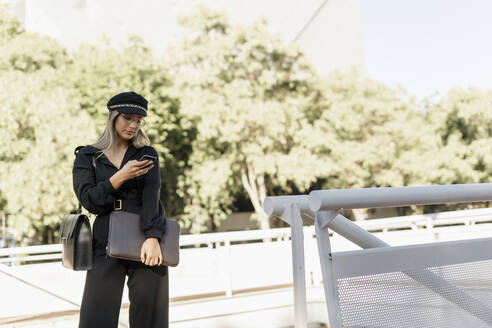 Young blond businesswoman wearing black hat and holding laptop bag and using smartphone - ERRF02078