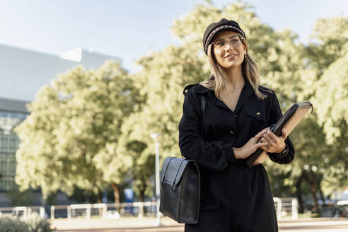 Junge blonde Geschäftsfrau mit schwarzer Matrosenmütze, die eine Laptoptasche hält und zur Seite schaut - ERRF02081