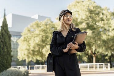 Junge blonde Geschäftsfrau mit schwarzer Seemannsmütze und Laptoptasche in der Hand - ERRF02082