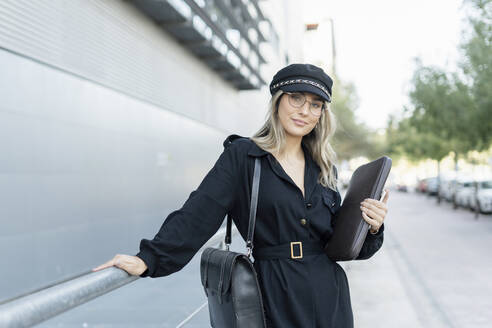 Junge blonde Geschäftsfrau mit schwarzer Seemannsmütze und Laptoptasche in der Hand - ERRF02084