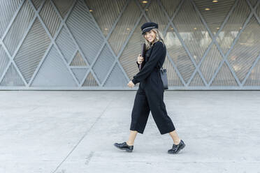 Junge blonde Geschäftsfrau mit schwarzer Matrosenmütze, Laptop-Tasche in der Hand, geht spazieren und schaut in die Kamera - ERRF02093