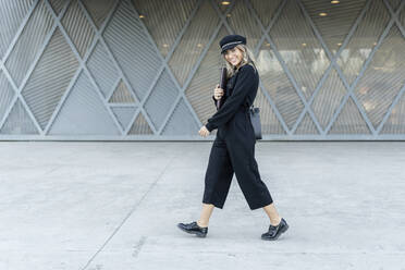 Young blond businesswoman wearing black sailor's cap, holding laptop bag, walking and looking at camera - ERRF02093