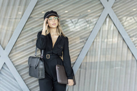 Junge blonde Geschäftsfrau mit schwarzem Hut und Laptoptasche in der Hand, Blick zur Seite, lizenzfreies Stockfoto