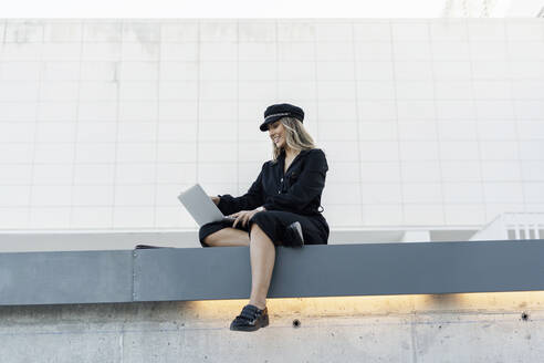 Junge blonde Geschäftsfrau mit schwarzer Matrosenmütze und Laptop, die an einer Wand sitzt - ERRF02097