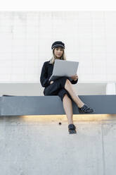 Junge blonde Geschäftsfrau mit schwarzer Matrosenmütze und Laptop, die an einer Wand sitzt - ERRF02098