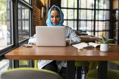 Businesswoman wearing turquoise hijab in a cafe and working, using laptop stock photo