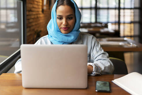 Geschäftsfrau mit türkisfarbenem Hidschab in einem Café bei der Arbeit, mit Laptop - ERRF02106