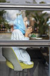 Junge Frau, die einen türkisfarbenen Hijab trägt und in einem Café Tee trinkt, schaut durch ein Fenster - ERRF02119