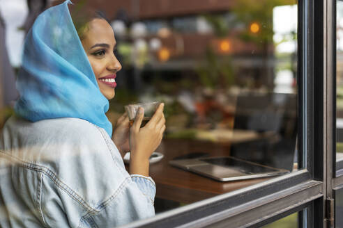 Junge Frau, die einen türkisfarbenen Hijab trägt und in einem Café Tee trinkt, schaut durch ein Fenster - ERRF02120