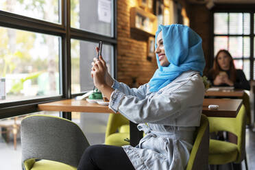 Junge Frau mit türkisfarbenem Hijab und Smartphone in einem Café - ERRF02133