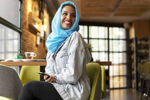 Junge Frau mit türkisfarbenem Hijab und Smartphone in einem Café - ERRF02137