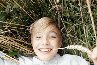 Porträt eines lächelnden blonden Jungen mit einer Haferähre im Mund, der in einem Haferfeld liegt - EYAF00691