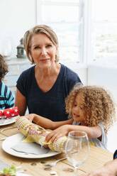 Woman with granddaughter playing with Christmas cracker at dining table - ISF22746