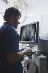 Junger männlicher Röntgenassistent, der eine Computertastatur benutzt, um eine Röntgenaufnahme des Beckens in der radiologischen Abteilung zu betrachten - ISF22789