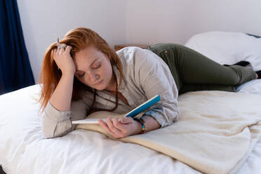 Young woman with red hair lying on bed reading her diary - ISF22799