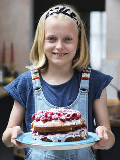Mädchen backt einen Kuchen, hält selbstgebackenen Kuchen mit Himbeeren in der Küche, Porträt - CUF53267
