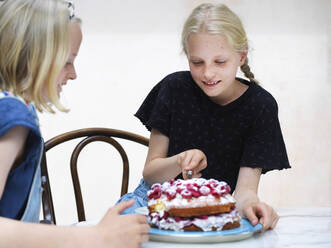 Mädchen und ihre Schwester schneiden ihren selbstgebackenen Kuchen mit frischen Himbeeren am Küchentisch an - CUF53269