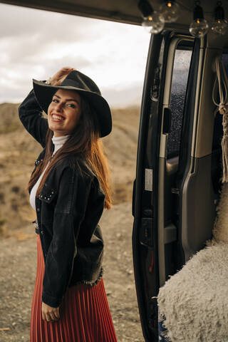 Porträt einer lächelnden jungen Frau in einer Wüstenlandschaft, die neben einem Wohnmobil steht, Almeria, Andalusien, Spanien, lizenzfreies Stockfoto