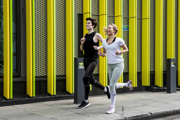 Two young female runners running along city sidewalk, full length - CUF53428