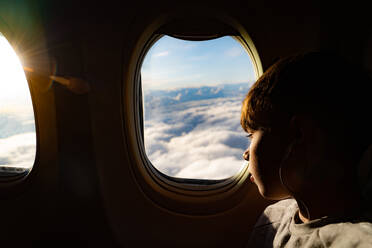 Teenage boy looking out through airplane window at clouds on airplane journey - CUF53529