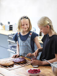 Mädchen und ihre Schwester backen einen Kuchen, streichen Marmelade auf den Kuchen am Küchentisch - CUF53532