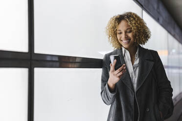 Lächelnde Frau, die mit ihrem Smartphone an einer Wand lehnt - AHSF01276