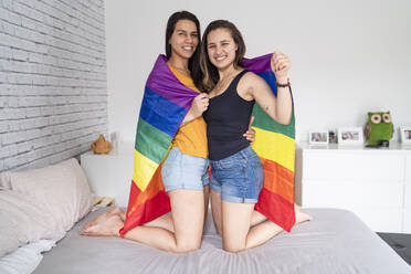 Porträt eines glücklichen lesbischen Paares, eingewickelt in eine Regenbogenflagge im Bett - FBAF00954