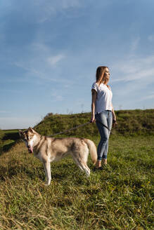 Junge Frau steht mit ihrem Hund auf einer Wiese bei Sonnenuntergang - MAUF03080