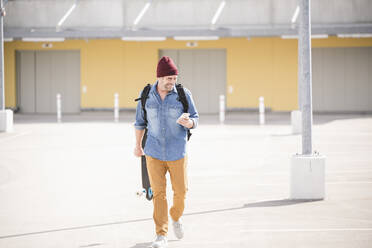 Casual mature man with smartphone and skateboard walking on parking deck - UUF19714