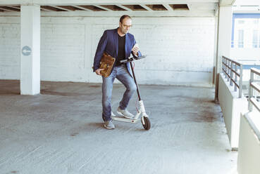 Mature businessman with e-scooter and smartphone in parking garage - UUF19716